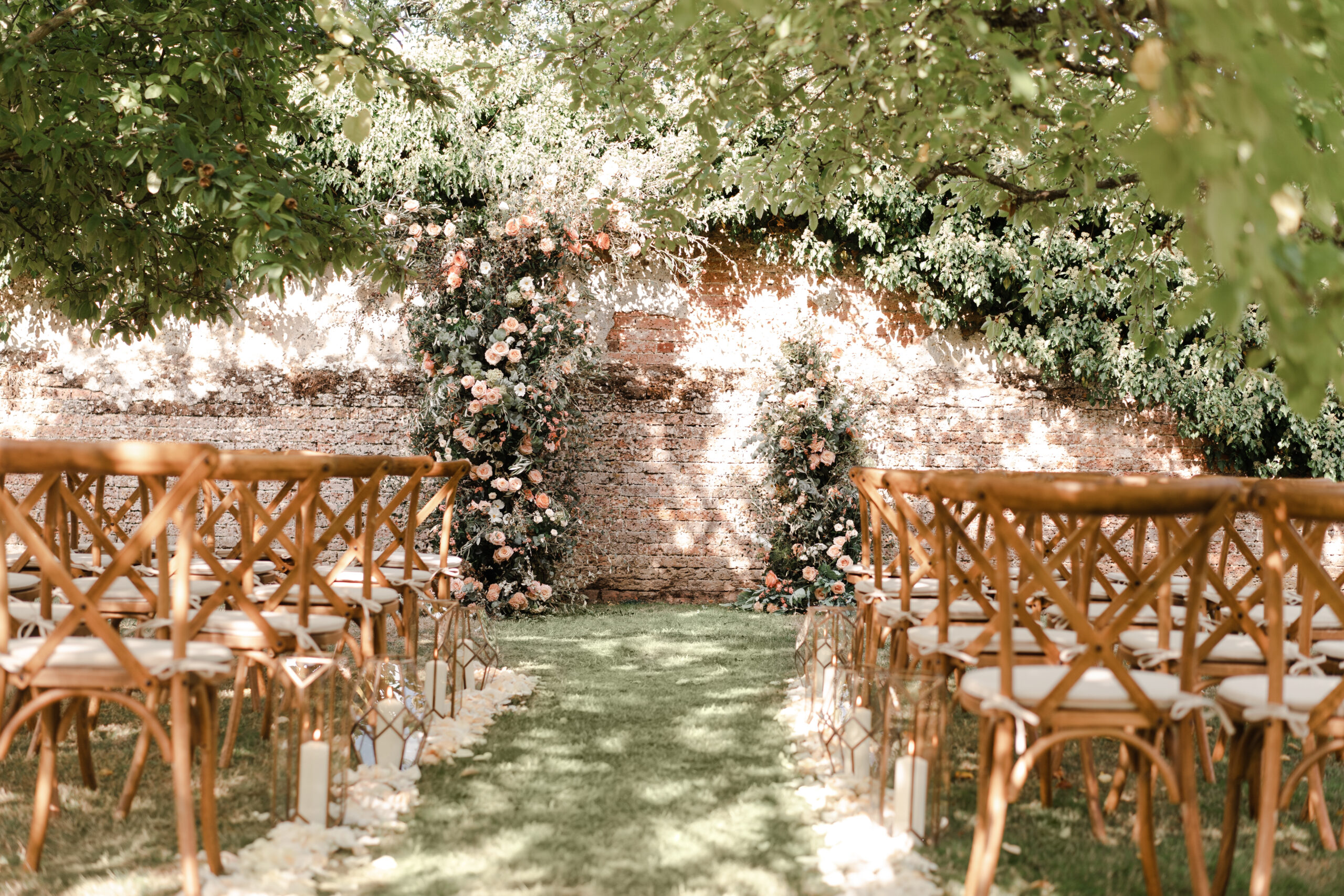 Four Seasons Hampshire Orchard Wedding Ceremony set up with petals and candles lining the aisle and a gorgeous coral and peach asymmetrical floral arch with whimsical greenery marking the end of the aisle