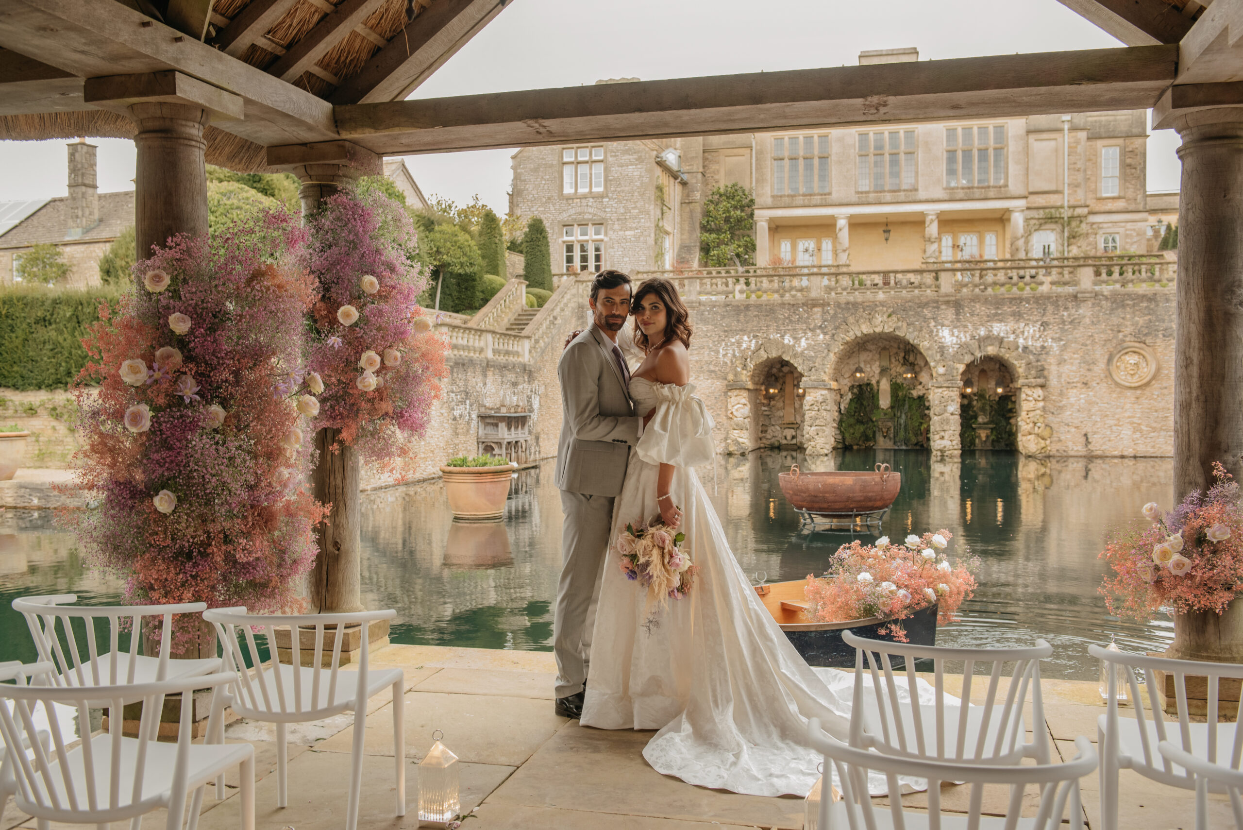 Wedding couple infront of the lake at Euridge manor. They stand on the boathouse surronded by sustainable flowers in pastel colours. Groom wears a grey Skopes lifecycle suit and bride wears a Emma Beumont bridal dress