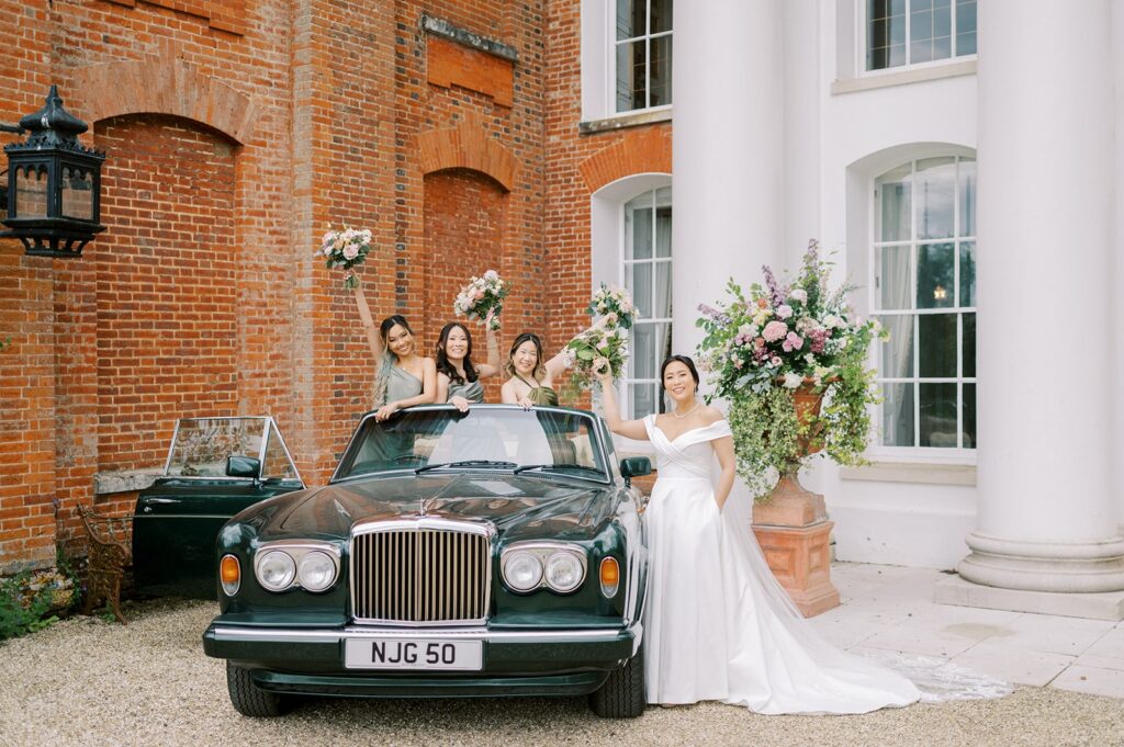 Amy and her bridesmaids celebrate on a classic car at their weekend wedding at Avington Park