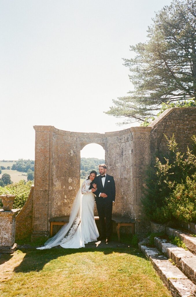 When it comes to how to choose your wedding photographer, do they also shoot film? Image is a film photograph of a couple standing next to an arch.