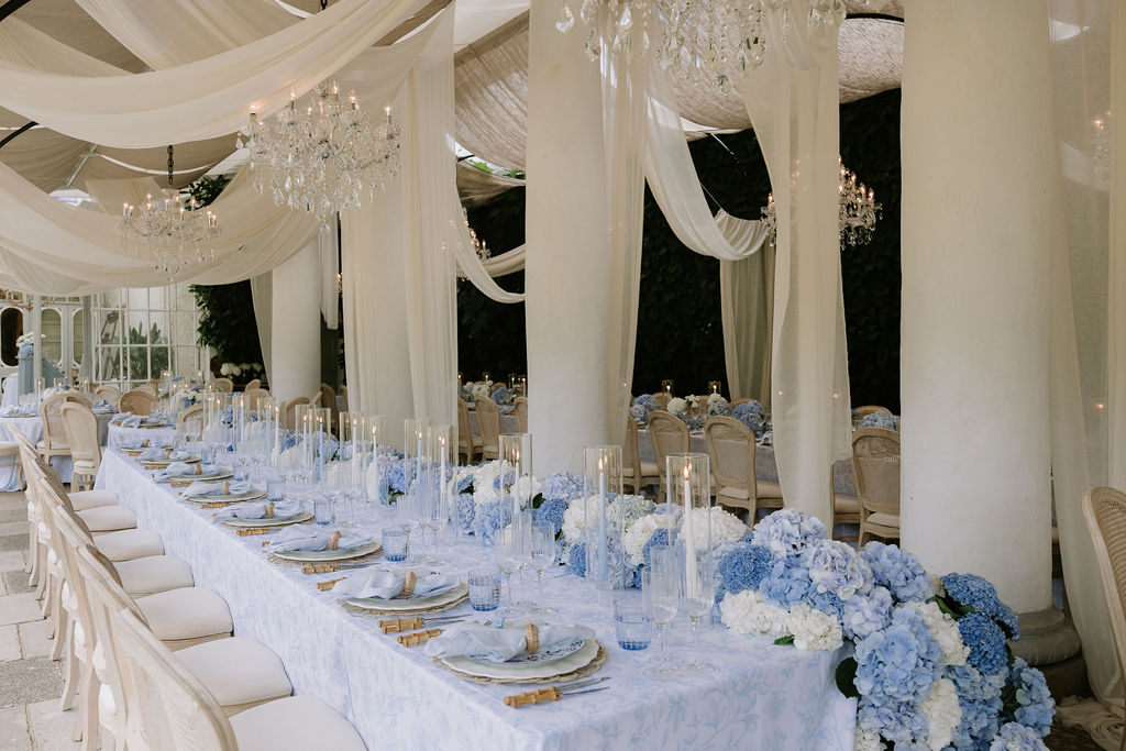 When it comes to how to choose your wedding photographer, consider their editing style. Image shows a table set for a wedding breakfast with blue hydrangeas and candles, and draping from the ceiling.